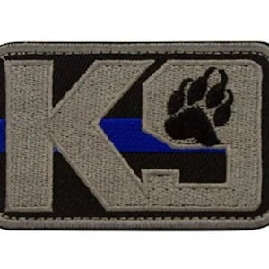 Peut inclure: Un écusson brodé gris et noir avec la lettre "K" et une empreinte de patte. L'écusson a une fine bande bleue au milieu.