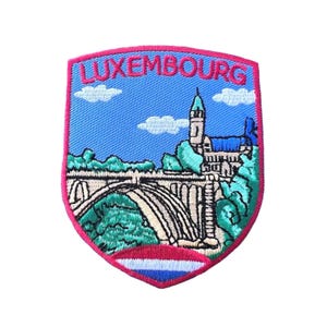 Puede incluir: Parche bordado con forma de escudo que muestra la palabra "LUXEMBOURG". El diseño incluye un puente, edificios y un cielo azul con nubes. El borde es rosa y la parte inferior tiene una franja roja, blanca y azul.