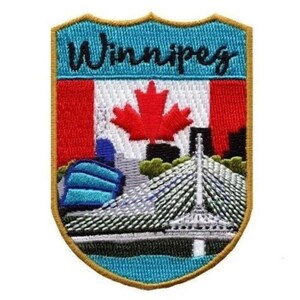 Op de afbeelding: Geborduurde patch in de vorm van een schild met een blauwe achtergrond en gouden rand. De patch toont een gestileerde stadsgezicht van Winnipeg, Canada, met de Canadese vlag en de tekst "Winnipeg" bovenaan.