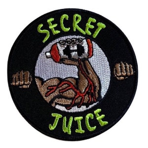 May include: A black and green embroidered patch with a white circle in the centre. The circle features a brown arm holding a red and white bottle with the text "SECRET JUICE" in green.