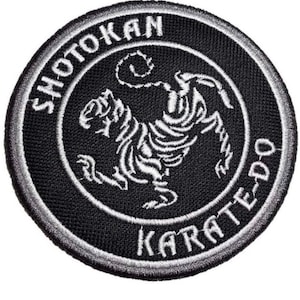 Puede incluir: Un parche bordado en blanco y negro con un tigre y el texto "Shotokan Karate-Do".