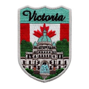 Pode incluir: Patch bordado com a palavra "Victoria" acima de uma bandeira canadense e uma ilustração detalhada de um edifício com uma cúpula e uma fonte. O patch tem uma borda branca e uma forma de escudo.
