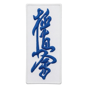 Op de afbeelding: Een rechthoekige witte patch met blauw geborduurde Japanse karakters. De karakters zijn gestileerd en verticaal gerangschikt. De patch heeft een witte rand en is ontworpen om op kleding of uitrusting te worden genaaid.