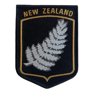 Może przedstawiać: Czarna i złota naszywka z napisem "NEW ZEALAND". Przedstawia srebrny wzór paproci. W kształcie tarczy, idealna do przyszycia do odzieży lub toreb.