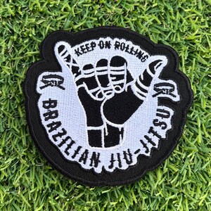 May include: Black and white embroidered patch with a hand gesture and the text "Keep on Rolling Brazilian Jiu-Jitsu".