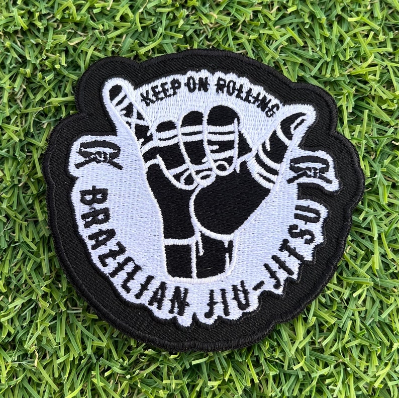 May include: Black and white embroidered patch with a hand gesture and the text "Keep on Rolling Brazilian Jiu-Jitsu".