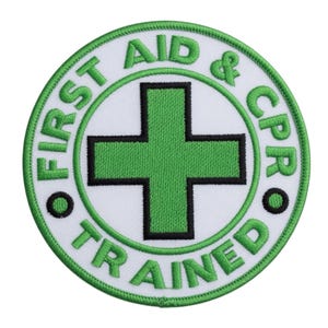 May include: A circular white patch with green and black embroidered text and a green cross symbol. The text reads "FIRST AID & CPR TRAINED" in a circular pattern around the cross.