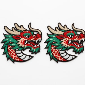 Könnte beinhalten: Zwei gestickte Drachenaufnäher. Jeder Aufnäher zeigt einen Drachenkopf mit roten, grünen und beigen Details. Das Maul des Drachen ist geöffnet und zeigt weiße Zähne. Auf weißem Hintergrund.