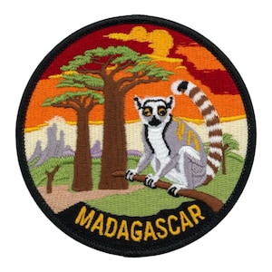 Pode incluir: Patch bordado circular com um lémure de cauda anelada empoleirado num galho, contra um cenário de pôr do sol e árvores baobá. A palavra "MADAGASCAR" é exibida em letras amarelas.
