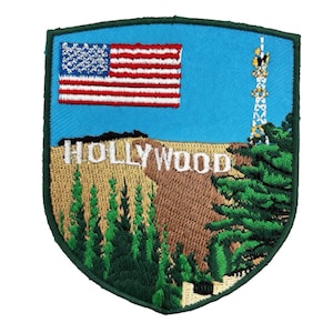 Op de afbeelding: Geborduurde patch met het Hollywood-teken, een Amerikaanse vlag en een radiotoren tegen een blauwe lucht. De patch heeft een schildvorm met een groene rand en toont een landschap met bomen en een bruine heuvel.