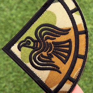 Op de afbeelding: Geborduurde patch met een gestileerd zwart vogelontwerp op een camouflage-achtergrond van groen, beige en cr&egrave;me. De patch heeft een zwarte rand en een opvallende vorm, ideaal voor het personaliseren van kleding of accessoires.