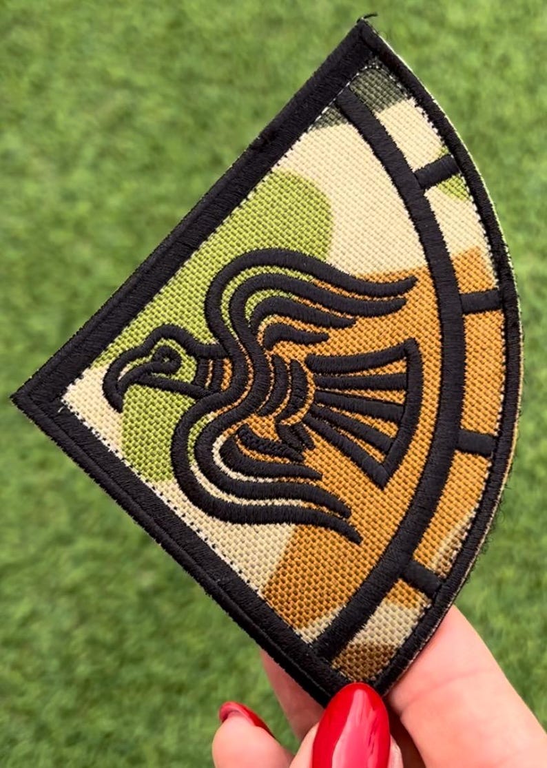 Op de afbeelding: Geborduurde patch met een gestileerd zwart vogelontwerp op een camouflage-achtergrond van groen, beige en cr&egrave;me. De patch heeft een zwarte rand en een opvallende vorm, ideaal voor het personaliseren van kleding of accessoires.
