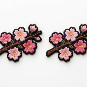 Pode incluir: Dois patches bordados com ramos de flores de cerejeira. Cada patch tem flores rosa com centros amarelos, folhas verdes e galhos marrons, todos delineados em preto. Os patches estão em um fundo branco.