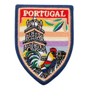 Op de afbeelding: Geborduurde patch met het woord "PORTUGAL" bovenaan, boven een gedetailleerde illustratie van een toren en een kleurrijke haan. De patch heeft een blauwe rand en een schildvorm, met een verscheidenheid aan kleuren.