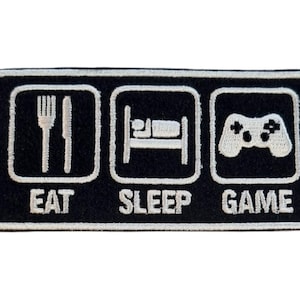 Könnte beinhalten: Schwarzer rechteckiger Aufnäher mit weißen gestickten Symbolen und Text. Der Aufnäher zeigt eine Gabel und ein Messer, ein Bett und einen Gamecontroller mit den Wörtern "EAT", "SLEEP" und "GAME" unter jedem Symbol.