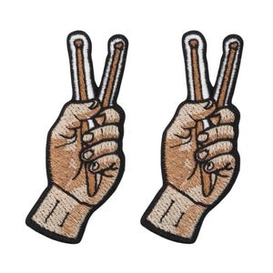 Op de afbeelding: Twee geborduurde patches met een hand die het vredesteken maakt. De handen zijn lichtbruin met zwarte contouren en details. De vredestekens zijn gemaakt met bruine en witte drumstokken.