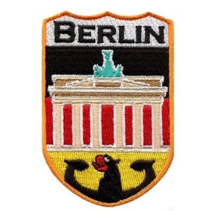 May include: A yellow, orange and black embroidered patch with the word "BERLIN" in white lettering. The patch features a stylised image of the Brandenburg Gate and a black bear.