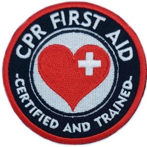 Può includere: Una toppa ricamata rossa, bianca e nera con il testo "CPR First Aid Certified and Trained" attorno a un cuore rosso con una croce bianca.