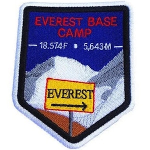 Könnte beinhalten: Gestickter Aufnäher mit blauem Hintergrund und einer weißen Bergkette. Der Text "EVEREST BASE CAMP" ist in Rot, und der Text "18,574F  5,643M" ist in Schwarz. Ein gelbes Schild mit dem Text "EVEREST" und einem Pfeil, der nach rechts zeigt, befindet sich im Vordergrund.