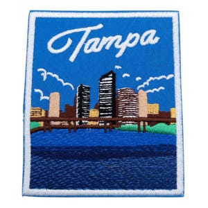 May include: Embroidered patch featuring the city of Tampa, Florida. The patch has a blue background with the city skyline, a bridge, and the word "Tampa" in white script. The patch is bordered in white.