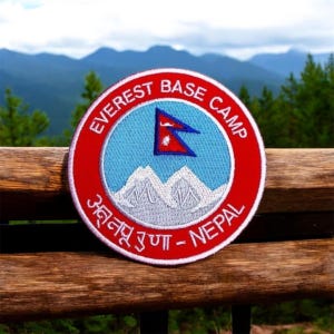 Pode incluir: Um remendo bordado em vermelho, branco e azul com o texto "Everest Base Camp" e "अन्नपूर्ण - Nepal". O remendo apresenta uma cordilheira estilizada e a bandeira do Nepal.