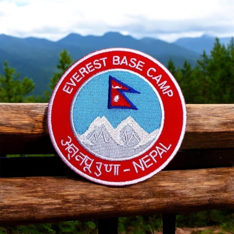 Pode incluir: Um remendo bordado em vermelho, branco e azul com o texto "Everest Base Camp" e "अन्नपूर्ण - Nepal". O remendo apresenta uma cordilheira estilizada e a bandeira do Nepal.