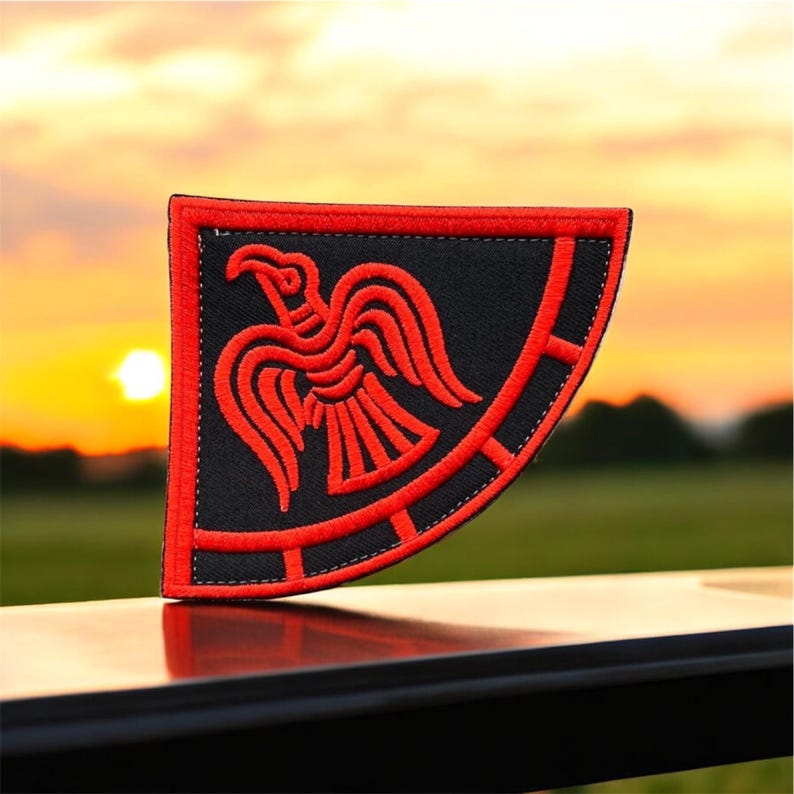 May include: A red embroidered Viking raven patch on a black background. The patch is shaped like a shield and has a circular design around the raven.