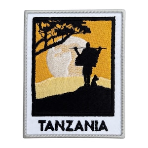 May include: Embroidered patch featuring a silhouette of a person carrying a load, a dog, and a tree against a yellow and white background. The word "TANZANIA" is printed in white block letters at the bottom. The patch has a white border.