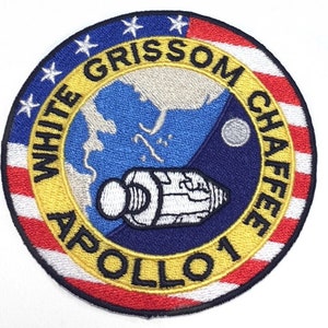Könnte beinhalten: Ein blauer und weißer gestickter Aufnäher mit goldener Umrandung und dem Text "WHITE GRISSOM CHAFFEE APOLLO 1" am Rand. Der Aufnäher zeigt ein stilisiertes Bild eines Raumschiffs vor dem Hintergrund der Erde und des Mondes.