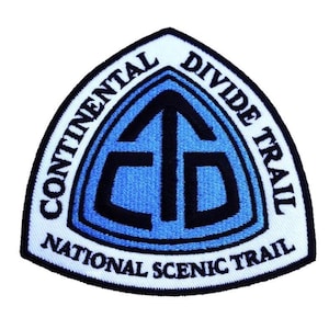 Könnte beinhalten: Ein weißer dreieckiger Patch mit dem Text "Continental Divide Trail National Scenic Trail" um den Umfang. Die Mitte des Patches zeigt einen blauen und schwarzen stilisierten Pfeil, der nach oben zeigt.