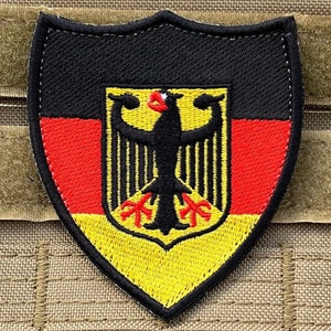Op de afbeelding: Een zwarte, rode en gele geborduurde patch in de vorm van een schild met het Duitse wapenschild, een zwarte adelaar met rode klauwen en snavel, op een gele achtergrond.