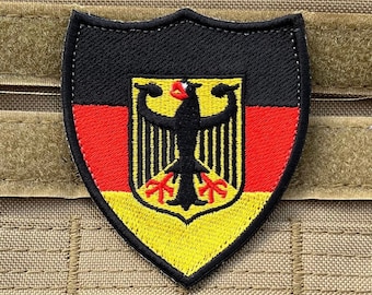 Könnte beinhalten: Ein schwarzer, roter und gelber gestickter Aufnäher in Form eines Schildes mit dem deutschen Bundeswappen, einem schwarzen Adler mit roten Krallen und Schnabel, auf gelbem Hintergrund.