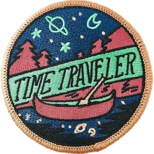 May include: A round embroidered patch with a dark blue background featuring a canoe, mountains, stars, and planets. The text "TIME TRAVELER" is written in green on a green banner.