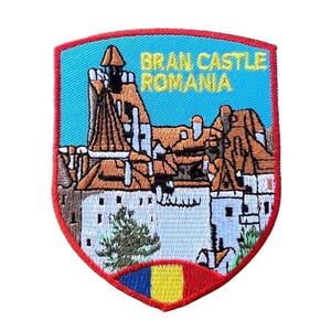 May include: An embroidered patch featuring a depiction of Bran Castle in Romania. The patch is shaped like a shield and has a red border. The castle is depicted in detail, with brown roofs and white walls. The text "Bran Castle Romania" is embroidered in yellow on a blue background.