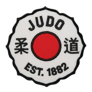 Könnte beinhalten: Ein weißer Aufnäher mit einem schwarzen, gezackten Rand. Er zeigt das Wort "JUDO" in schwarzen Blockbuchstaben, einen roten Kreis in der Mitte, japanische Schriftzeichen und den Text "EST. 1882" in Schwarz.