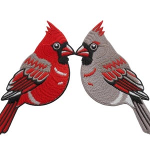 May include: Two embroidered bird patches featuring a bright red cardinal and a grey cardinal facing each other. Both birds have black facial markings around their beaks, crested heads, and detailed wing feathers in red, white, and black thread.