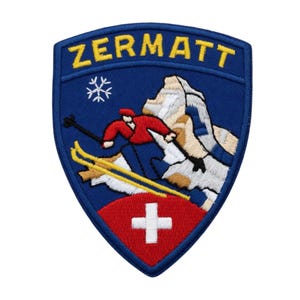 Peut inclure: Écusson brodé en forme de bouclier bleu avec le mot "ZERMATT" en jaune. Un skieur en rouge dévale une montagne, avec un drapeau suisse en bas. Un flocon de neige est au-dessus du skieur.
