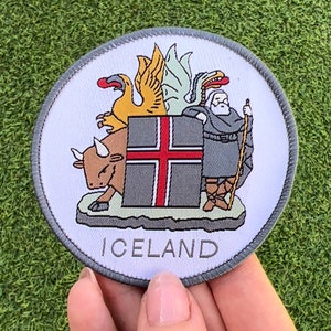 Pode incluir: Um remendo bordado branco e cinza com o brasão de armas da Islândia. O brasão de armas apresenta um escudo com uma cruz vermelha sobre um fundo branco, um touro, um grifo e um bispo segurando um cajado. O remendo é rodeado por uma borda cinza e a palavra "ICELAND" está impressa abaixo do brasão de armas.