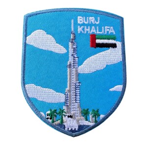 Pode incluir: Patch bordado azul claro em forma de escudo com o Burj Khalifa. O patch tem uma borda e fundo azul claro, com nuvens brancas e palmeiras. O texto "BURJ KHALIFA" e a bandeira dos Emirados Árabes Unidos são visíveis.