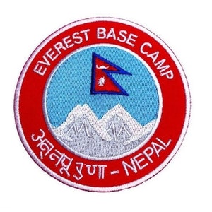 May include: An embroidered patch in red and white with the text "Everest Base Camp" and "अन्नपूर्ण - Nepal" in Nepali. The patch features a white mountain range and a blue and red flag of Nepal.