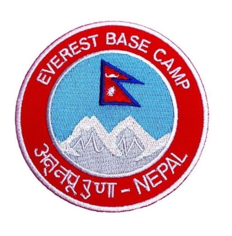 Pode incluir: Um remendo bordado em vermelho e branco com o texto "Everest Base Camp" e "अन्नपूर्ण - Nepal" em nepal&ecirc;s. O remendo apresenta uma cordilheira de montanhas brancas e uma bandeira do Nepal azul e vermelha.
