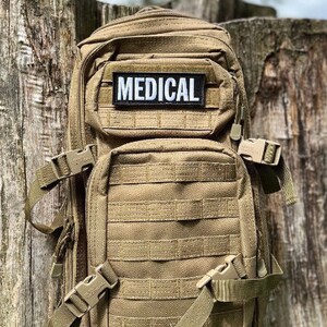 May include: A tan tactical backpack with multiple compartments and MOLLE webbing. A black and white patch with the word "MEDICAL" is centered on the front. Features adjustable straps and secure buckles for carrying medical supplies or gear.