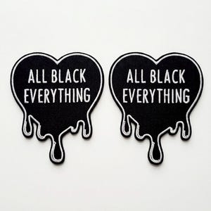 Könnte beinhalten: Zwei schwarze herzförmige Aufnäher mit weiß gestickten Rändern und dem Schriftzug "ALL BLACK EVERYTHING" in weißen Blockbuchstaben. Der Boden jedes Herzens hat ein tropfendes Design.