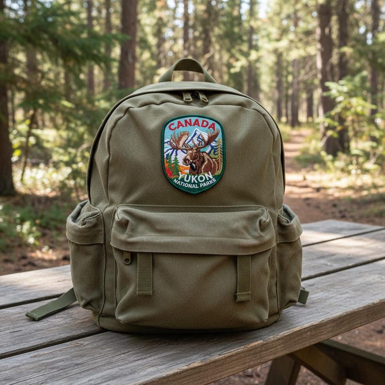 May include: An olive green backpack featuring a large embroidered patch on the front pocket. The patch displays a moose, mountains, and trees with the text CANADA YUKON NATIONAL PARKS. The bag has a main compartment, a front pocket, and side pockets.