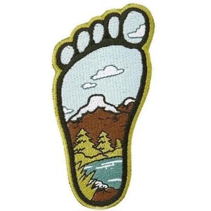 Może przedstawiać: Naszywka haftowana w kształcie odcisku stopy z górskim krajobrazem w środku. Krajobraz zawiera jezioro, drzewa i górę ze śnieżnym szczytem. Naszywka jest obwiedziona żółtym kolorem.