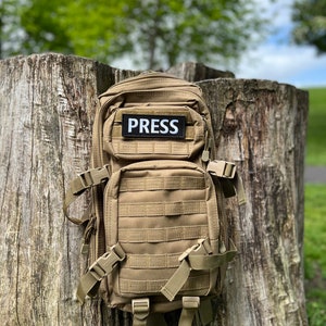 Peut inclure: Un sac &agrave; dos tactique beige avec un patch noir sur lequel est inscrit "PRESS". Le sac &agrave; dos est pos&eacute; sur une souche d'arbre.