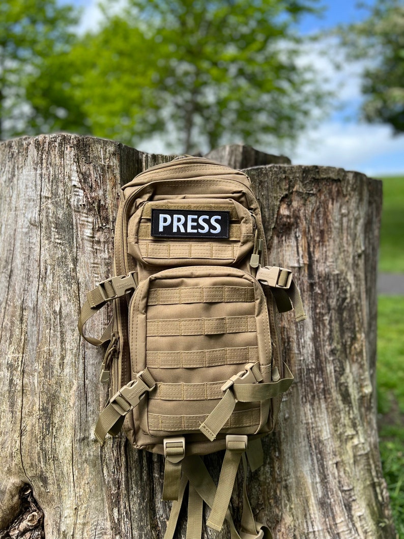 Peut inclure: Un sac &agrave; dos tactique beige avec un patch noir sur lequel est inscrit "PRESS". Le sac &agrave; dos est pos&eacute; sur une souche d'arbre.