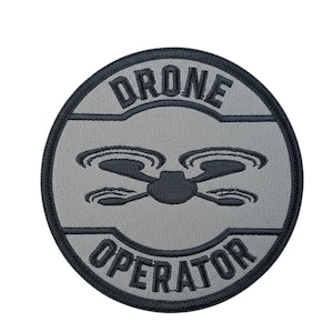 May include: A circular patch with a gray background and black border. The patch features the words "DRONE OPERATOR" in a curved design, with a drone graphic in the center. The design is embroidered.