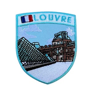 May include: A light blue embroidered patch featuring the Louvre Museum in Paris. The patch has a shield shape with a French flag and the word "LOUVRE" in blue letters. The image depicts the glass pyramid and the museum building.