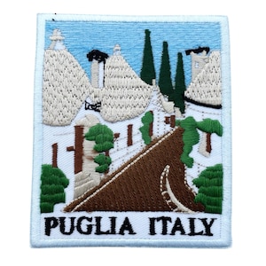 Puede incluir: Parche bordado que representa la región de Apulia, Italia. El diseño incluye edificios blancos con tejados cónicos, árboles verdes y el texto "PUGLIA ITALY" en la parte inferior. El parche tiene un borde azul claro y es adecuado para chaquetas, bolsos u otros artículos de tela.